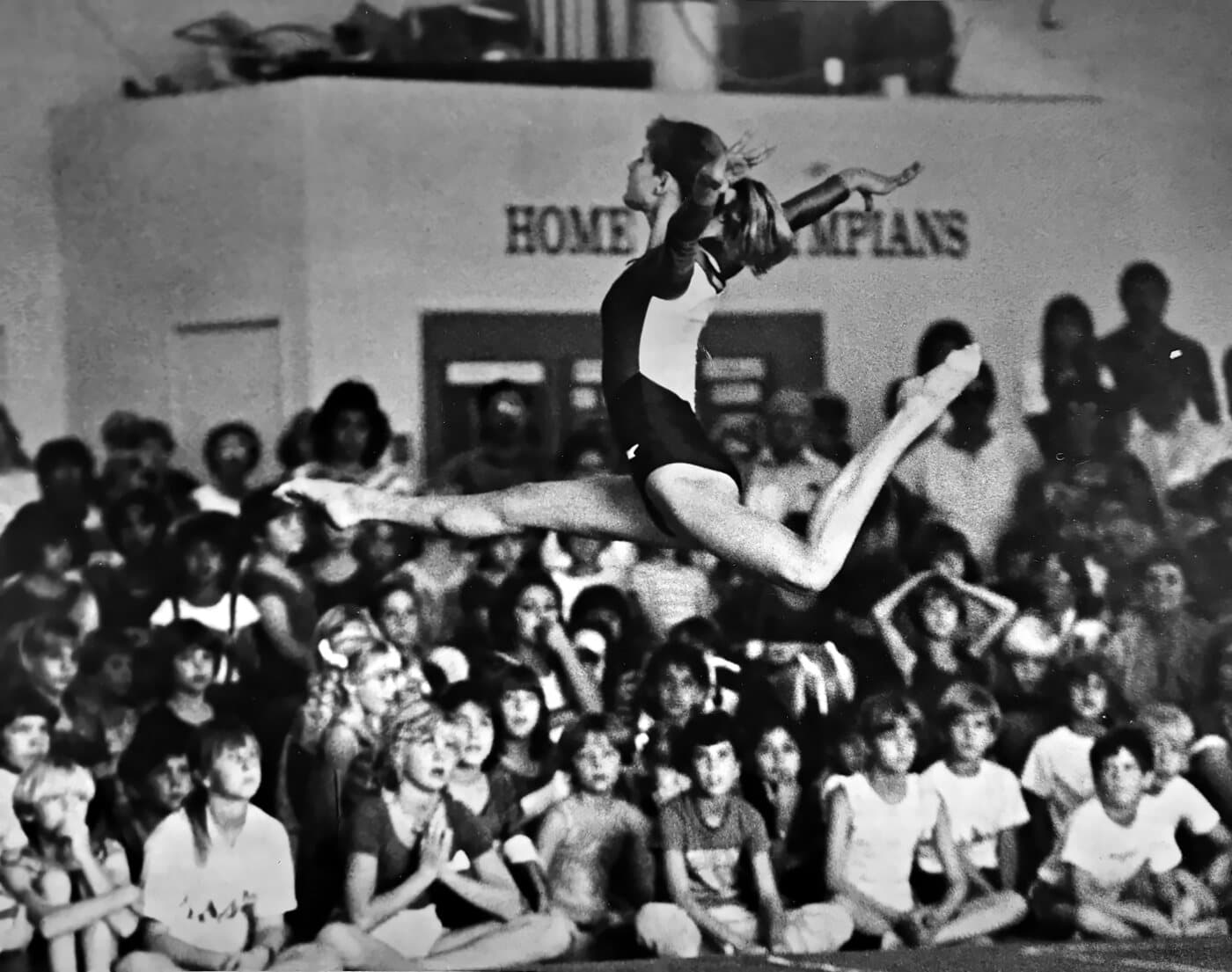 Kristine mid-leap in a full split above a packed gym floor, spectators seated below